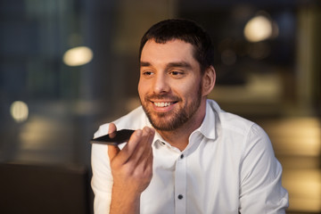business, deadline and technology concept - happy businessman using voice command recorder on smartphone at night office