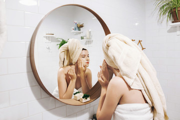 Young happy woman in towel making facial massage with  organic face scrub and looking at mirror in...