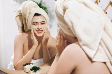 Young happy woman in towel making facial massage with  organic face scrub and looking at mirror in...