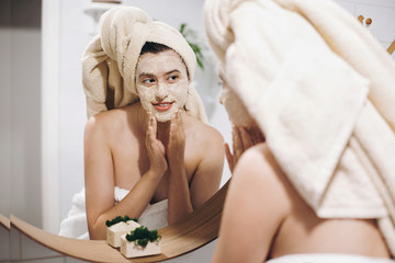 Obraz na płótnie Canvas Young happy woman in towel applying organic face mask and looking at round mirror in stylish bathroom. Girl making facial massage with scrub, peeling and cleaning skin on face. Skin Care