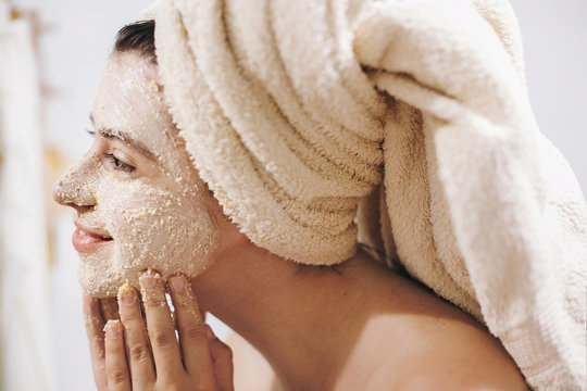 Skin Care concept. Young happy woman in towel making facial massage with organic face scrub close up in stylish bathroom. Girl applying scrub cream, peeling and cleaning skin.