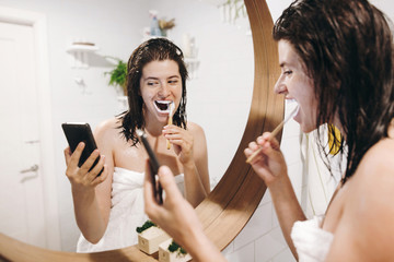 Young happy woman in white towel brushing teeth and looking at smartphone in bathroom with mirror. Slim sexy woman with natural skin and wet hair daily routine after shower. Social media affect