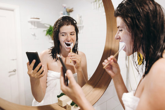 Young happy woman in white towel brushing teeth and looking at smartphone in bathroom with mirror. Slim sexy woman with natural skin and wet hair daily routine after shower. Social media affect