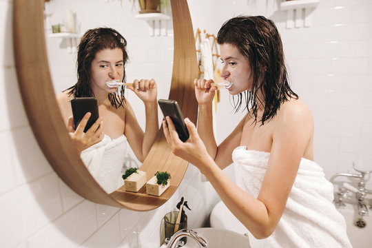 Social media affect. Young happy woman in white towel brushing teeth and looking at phone screen in stylish bathroom at round mirror. Slim sexy woman daily routine after shower.