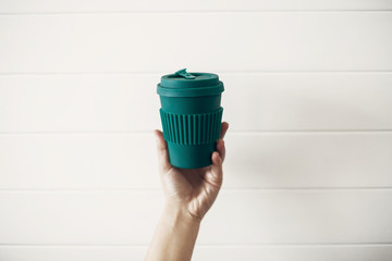 Hand holding stylish reusable eco coffee cup on white wooden background. Green Cup from natural ...