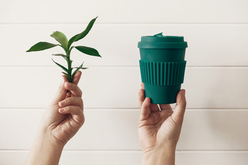 Hands holding stylish reusable eco coffee cup and green bamboo leaves on white wooden background....