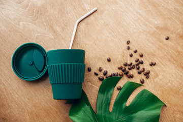 Zero waste concept, flat lay. Stylish reusable eco coffee cup and metallic steel straws on wooden background with green monstera leaf. Ban single use plastic. Sustainable lifestyle