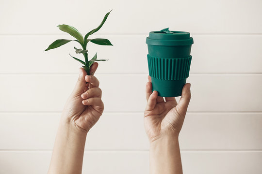 Zero waste concept, sustainable lifestyle. Hands holding stylish reusable eco coffee cup and green bamboo leaves on white wooden background. Ban single use plastic. Make a choice.