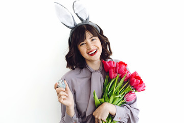 Beautiful girl smiling with bunny ears, holding easter egg and tulips on white background indoors, space for text. Easter hunt concept,spring  seasonal greetings.