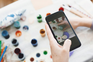 Hands holding phone and taking photo of stylish easter flat lay of painting egg on rustic table with paint,brushes, bird toy. Easter workshop.Decorating easter egg, holiday preparations.