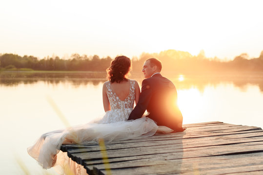 young beautiful bride and groom resting on the pier near the river at sunset