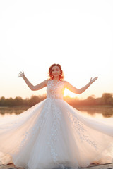 young beautiful bride in white dress on wooden pier near water at sunset