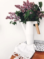 Beautiful lilac flowers in white stylish watering can on white background indoors. Modern decor design, purple lilac in metal funnel vase with bamboo handle. Hello spring