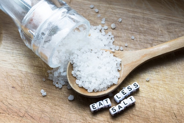 Eat less salt message written near wooden spoon full of granulated salt and shaker on rustic...