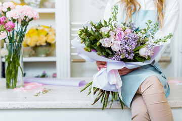 cropped view of female florist sitting on counter and holding bouquet with lilac and roses in flower shop