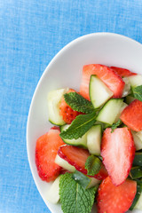 Raw fruit detox salad with cucumbers strawberries apples fresh mint in crystal bowl on blue table cloth background. Vegan plant base diet healthy food concept. Copy space