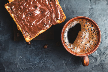 Cup with hot coffee and toast with chocolate hazelnut cream on dark background, top view