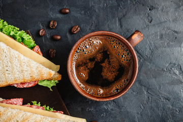 Cup with hot coffee and sandwich with grilled toast, salami sausage, salad lettuce and cheese on dark background, top view