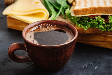 Cup with hot coffee and sandwich with grilled toast, salami sausage, salad lettuce and cheese on dark background