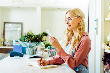 beautiful female flower shop owner in glasses writing in notebook and using smartphone