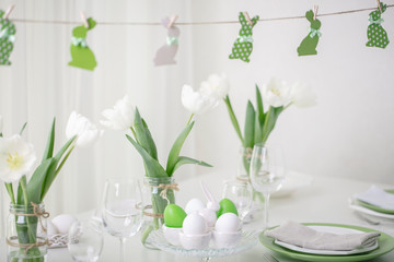 Decor and table setting of the Easter table with white tulips and dishes of green and white color. Easter decor in the form of Easter bunnies  green color with white polka dots.