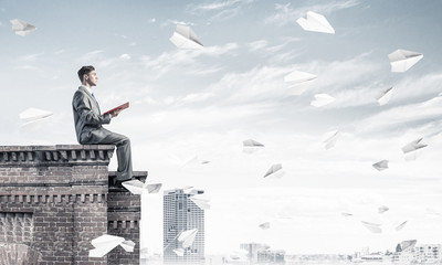 Man on roof edge reading book and cityscape at background