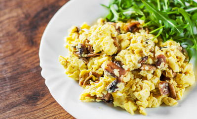 scrambled eggs with mushrooms and arugula salad in white plate on wooden table background