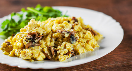 scrambled eggs with mushrooms and arugula salad in white plate on wooden table background