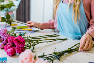 cropped view of female florist sitting at table with pink roses and carnations while arranging bouquet