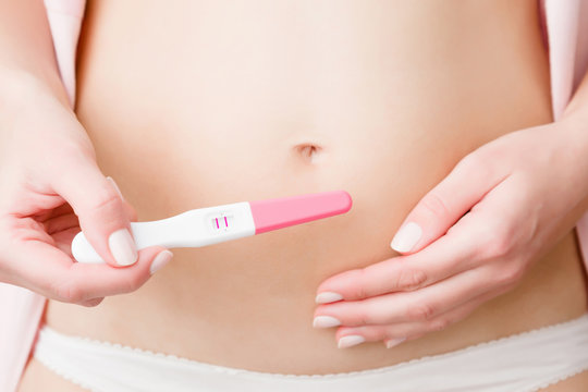 Young woman's hand holding pregnancy test with two stripes on naked belly. Positive result. Closeup. Front view. 