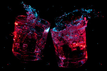 Big splashes of liquid glowing in colorful lights in two rocks glasses clinking in toast isolated on a black background