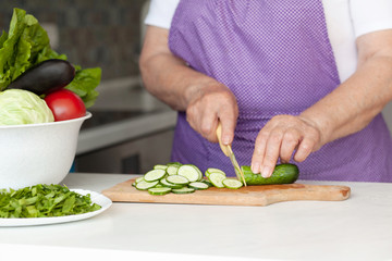 An old woman cutting fresh vegetables in the kitchen. Healthy eating. Preparation of salad. 