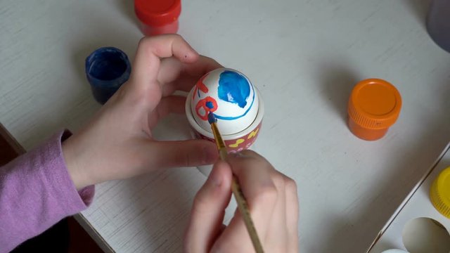 The child is preparing for the holiday and draws on the Easter egg symbols of blue and red paint, sitting at the table. Close up. The view from the top. 4K. 25 fps
