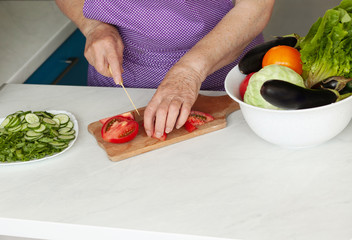 An old woman cutting fresh vegetables in the kitchen. Healthy eating. Preparation of salad. 