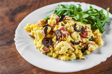 scrambled eggs with ham, olive and arugula salad in white plate on wooden table background