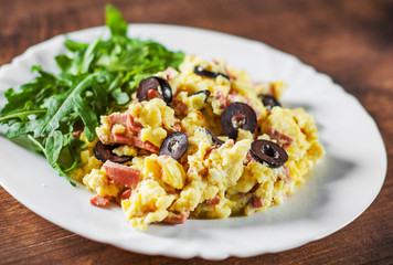 scrambled eggs with ham, olive and arugula salad in white plate on wooden table background