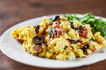 scrambled eggs with ham, olive and arugula salad in white plate on wooden table background