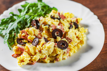 scrambled eggs with ham, olive and arugula salad in white plate on wooden table background