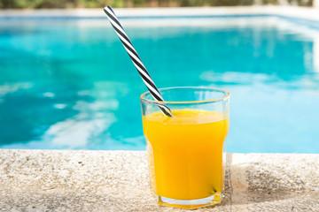Glass of freshly pressed tropical fruits orange juice with striped straw standing on deck of swimming pool. Bright sunlight. Summer vacation relaxation travel luxury escape. Lifestyle