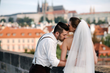 Naklejka premium Back view of young romantic wedding couple standing on the bridge and enjoying beautiful view of the city Prague, Czech Republic. Groom kissing bride
