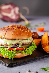 American burger with meat patty, tomato, cucumber, green sauce. Sauce and for french fries on a black stone slate board. Dish in a fast food restaurant. Copy space