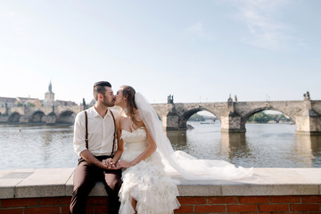 Young romantic wedding couple sitting on the bridge with the beautiful city view and kissing. Charles bridge, Prague, Czech Republic