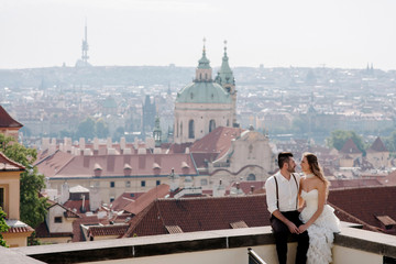Young romantic couple of groom and bride sitting on the rooftop and enjoying beautiful view of the city Prague, Czech Republic