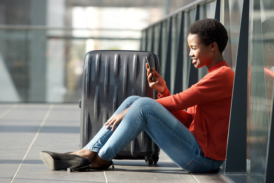 happy young african american woman sitting on floor in airport terminal with luggage and looking at mobile phone
