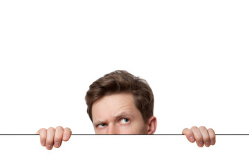 Young man with surprised eyes peeking out from behind billboard paper poster. Man peeking out from the edge and looking at camera isolated on a white background