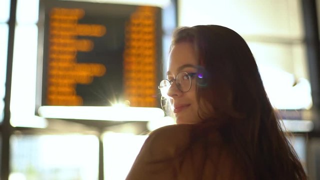 Slow motion rear-view behind charming european girl study departure board airport, train station, searching flight, sun shining camera, turning back smiling silly cute, excited travel foreign country
