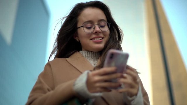 Slow motion low-angle happy charismatic european brunette wearing glasses, scyscraper urban background, looking smartphone, surprised silly funny message received, laughing looking mobile display