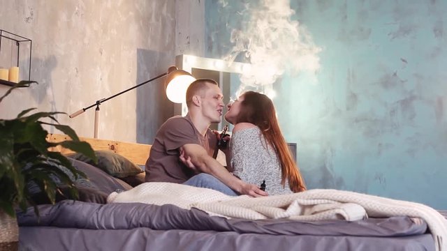 A loving couple on a bed smokes electronic cigarettes.