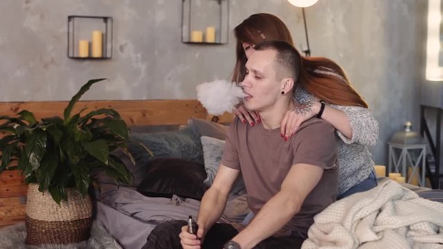A loving couple on a bed smokes electronic cigarettes.