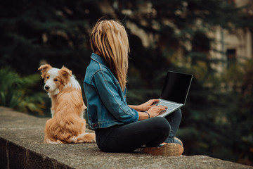 blonde Girl in cool jeans jacket checking something on her laptop, while sitting in the park with her dog